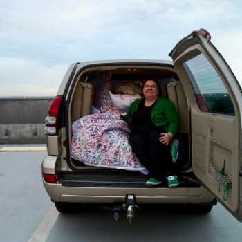 Woman sitting in the back of an open SUV filled with bedding, smiling while wearing a green cardigan and sneakers in a rooftop car park.